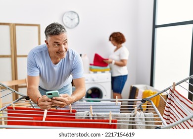 Middle Age Man And Woman Couple Using Smartphone Hanging Clothes At Laundry