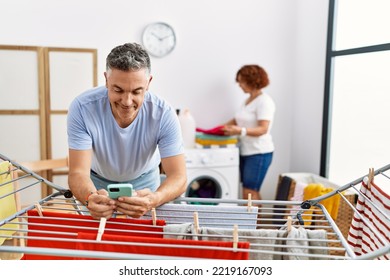 Middle Age Man And Woman Couple Using Smartphone Hanging Clothes At Laundry