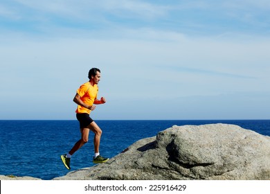 Middle Age Man At Workout Outdoors Jogging Along The Sea At Sunny Day, Male Jogger In Fluorescent T-shirt Runs Over Sea Rocks At Morning Training Outdoors