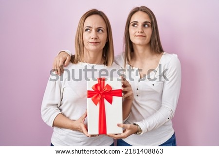 Middle age mother and young daughter holding mothers day gift smiling looking to the side and staring away thinking. 