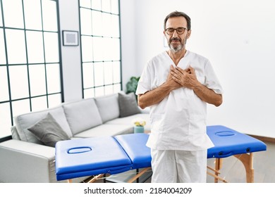 Middle Age Physiotherapy Man Working At Home Smiling With Hands On Chest With Closed Eyes And Grateful Gesture On Face. Health Concept. 