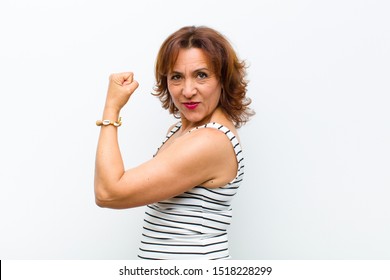 middle age pretty woman feeling happy, satisfied and powerful, flexing fit and muscular biceps, looking strong after the gym against white wall