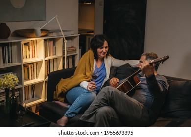 Middle Age Romantic Couple Drinking Wine While Sitting On Sofa At Home. Man Playing Acoustic Guitar