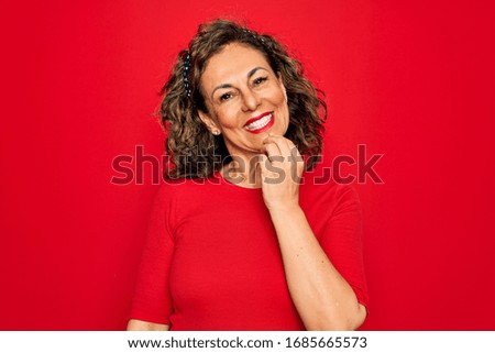 Middle age senior brunette woman wearing casual t-shirt standing over red background looking confident at the camera with smile with crossed arms and hand raised on chin. Thinking positive.