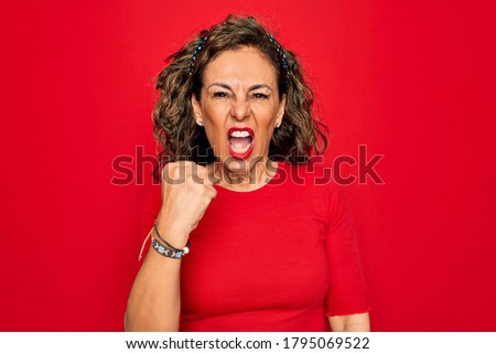 Middle age senior brunette woman wearing casual t-shirt standing over red background angry and mad raising fist frustrated and furious while shouting with anger. Rage and aggressive concept.