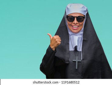 Middle age senior catholic nun woman wearing sunglasses over isolated background smiling with happy face looking and pointing to the side with thumb up.