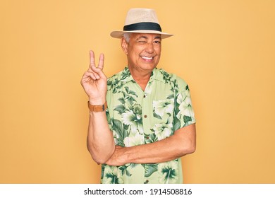 Middle Age Senior Grey-haired Man Wearing Summer Hat And Floral Shirt On Beach Vacation Smiling With Happy Face Winking At The Camera Doing Victory Sign. Number Two.