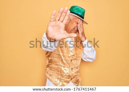 Middle age senior grey-haired man wearing Brazilian carnival custome over yellow background covering eyes with hands and doing stop gesture with sad and fear expression. Embarrassed and negative