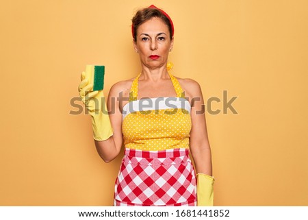 Middle age senior housewife pin up woman wearing 50s style retro dress using cleaner scrub with a confident expression on smart face thinking serious