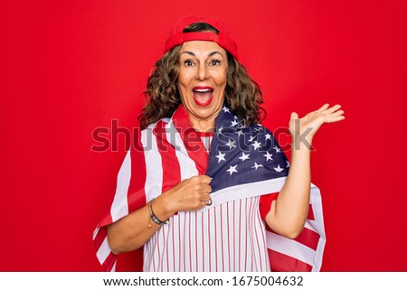 Middle age senior patriotic woman wearing baseball equipment holding USA flag very happy and excited, winner expression celebrating victory screaming with big smile and raised hands