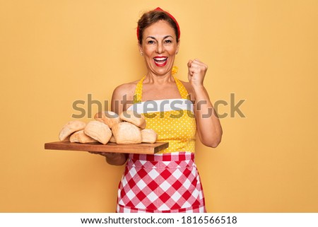 Middle age senior pin up housewife woman wearing 50s style retro dress and apron cooking bread screaming proud and celebrating victory and success very excited, cheering emotion