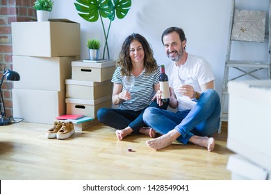 Middle age senior romantic couple in love sitting on the apartment floor with boxes around, celebrating drinking a glass of wine smiling happy for moving to a new home
