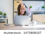 Middle age senior woman sitting at the table at home working using computer laptop surprised with hand on head for mistake, remember error. Forgot, bad memory concept.