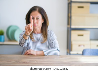 Middle age senior woman sitting at the table at home Pointing with finger up and angry expression, showing no gesture