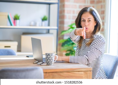 Middle age senior woman sitting at the table at home working using computer laptop pointing with finger to the camera and to you, hand sign, positive and confident gesture from the front