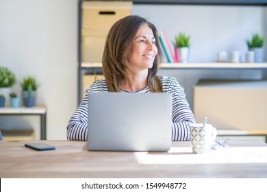 Middle Age Senior Woman Sitting At The Table At Home Working Using Computer Laptop Looking Away To Side With Smile On Face, Natural Expression. Laughing Confident.