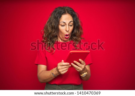 Middle age senior woman using touchpad tablet over red isolated background scared in shock with a surprise face, afraid and excited with fear expression