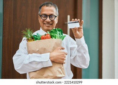 Middle Age Southeast Asian Man Smiling Holding A Bag Of Fresh Groceries Standing By Home Door With Credit Card