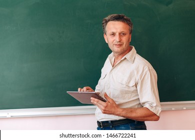 Middle Age Teacher Next The Blackboard Holding An Ipad And Explain A Lesson.