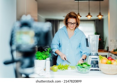 Middle Age Woman Is Blogging For Her Wellness Lifestyle Channel About Healthy Balanced Living In The Kitchen Of Her Home. Creating Video Content For Social Media With Camera On A Tripod.