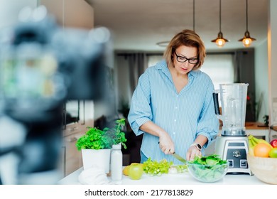 Middle Age Woman Is Blogging For Her Wellness Lifestyle Channel About Healthy Balanced Living In The Kitchen Of Her Home. Creating Video Content For Social Media With Camera On A Tripod