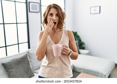 Middle Age Woman Drinking A Cup Coffee At Home Afraid And Shocked, Surprise And Amazed Expression With Hands On Face 