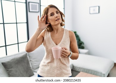 Middle Age Woman Drinking A Cup Coffee At Home Smiling With Hand Over Ear Listening An Hearing To Rumor Or Gossip. Deafness Concept. 