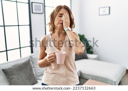 Middle age woman drinking a cup coffee at home yawning tired covering half face, eye and mouth with hand. face hurts in pain. 