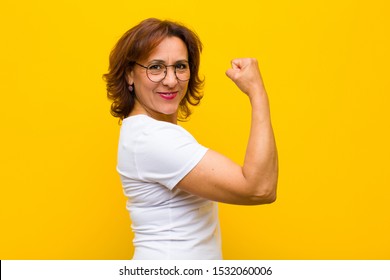 middle age woman feeling happy, satisfied and powerful, flexing fit and muscular biceps, looking strong after the gym against yellow wall