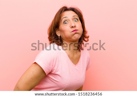 middle age woman with a goofy, crazy, surprised expression, puffing cheeks, feeling stuffed, fat and full of food against pink wall
