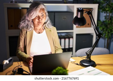 Middle Age Woman With Grey Hair Working Using Computer Laptop Late At Night Puffing Cheeks With Funny Face. Mouth Inflated With Air, Crazy Expression. 