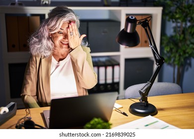 Middle Age Woman With Grey Hair Working Using Computer Laptop Late At Night Covering One Eye With Hand, Confident Smile On Face And Surprise Emotion. 