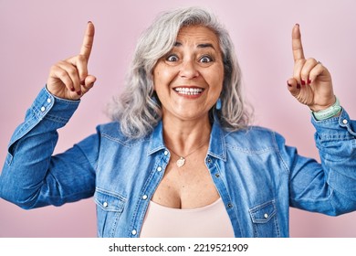 Middle Age Woman With Grey Hair Standing Over Pink Background Smiling Amazed And Surprised And Pointing Up With Fingers And Raised Arms. 