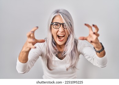 Middle age woman with grey hair standing over white background shouting frustrated with rage, hands trying to strangle, yelling mad 
