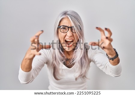 Middle age woman with grey hair standing over white background shouting frustrated with rage, hands trying to strangle, yelling mad 