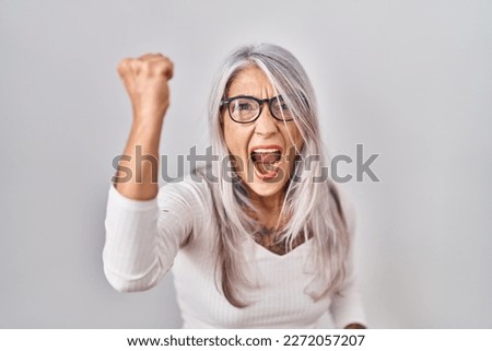 Middle age woman with grey hair standing over white background angry and mad raising fist frustrated and furious while shouting with anger. rage and aggressive concept. 