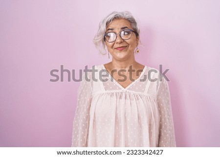Middle age woman with grey hair standing over pink background smiling looking to the side and staring away thinking. 