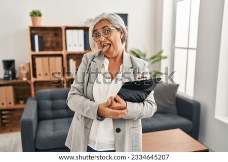 Middle age woman with grey hair at consultation office sticking tongue out happy with funny expression. emotion concept. 