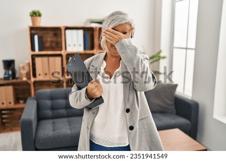 Middle age woman with grey hair at consultation office covering eyes with hand, looking serious and sad. sightless, hiding and rejection concept 