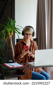 Middle Age Woman In Headphones And With Mobile Phone Woking With Laptop Computer At Home