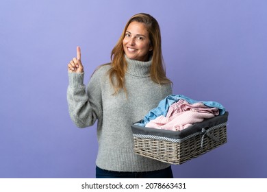 Middle age woman holding a clothes basket isolated on purple background showing and lifting a finger in sign of the best
