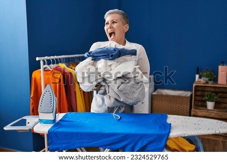 Middle age woman ironing clothes at home angry and mad screaming frustrated and furious, shouting with anger. rage and aggressive concept. 