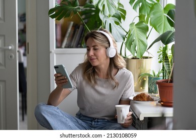 Middle age woman listening to music wear wireless headphones, using mobile smart phone chatting in social networks, sitting next to window drinking tea of coffee in the morning at home. Time to relax  - Powered by Shutterstock