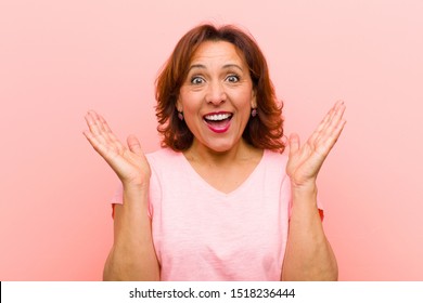 Middle Age Woman Looking Happy And Excited, Shocked With An Unexpected Surprise With Both Hands Open Next To Face Against Pink Wall