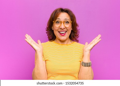 Middle Age Woman Looking Happy And Excited, Shocked With An Unexpected Surprise With Both Hands Open Next To Face Against Purple Wall