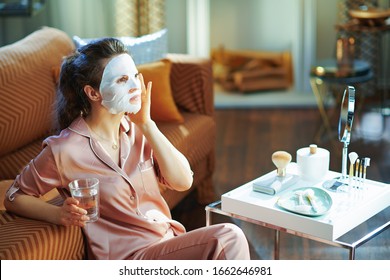 middle age woman in pajamas with white sheet facial mask on face and glass of water in the modern living room in sunny winter day.