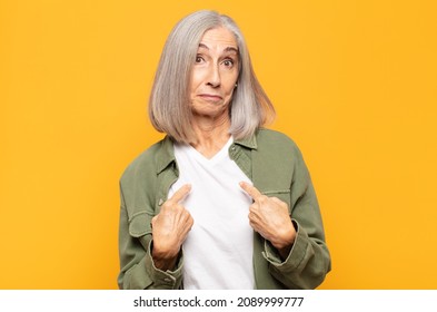 Middle Age Woman Pointing To Self With A Confused And Quizzical Look, Shocked And Surprised To Be Chosen