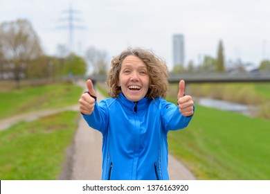 Middle age woman in running jacket showing thumbs up with both hands, standing outdoors in the park and smiling, looking at camera. Half-length front portrait