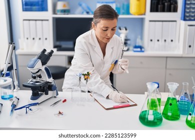 Middle age woman scientist holding test tube with plant writing on clipboard at laboratory - Powered by Shutterstock