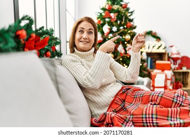 Middle Age Woman Sitting On The Sofa By Christmas Tree Smiling And Looking At The Camera Pointing With Two Hands And Fingers To The Side. 